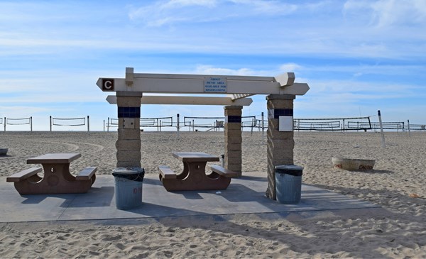 Newland Ramada C sits on a paved path (wheelchair accessible). There are two picnic tables and a fire ring adjacent to the area. Volleyball courts line the sand several yards behind the picnic area.